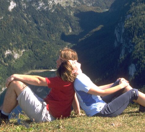 A couple sits back to back in a high meadow, with a view of the Alpine panorama in the Salzkammergut region