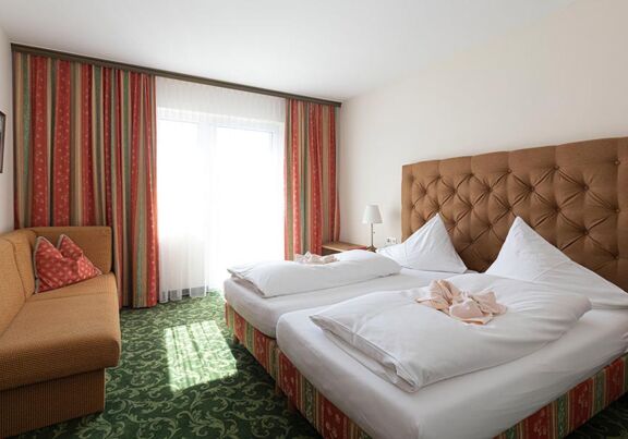 Ein Hotelzimmer mit grünem Teppich und einem Doppelbett. Gegenüber ist ein Sofa und daneben ein Fenster mit Vorhängen.
