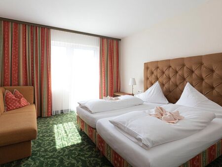 Ein Hotelzimmer mit grünem Teppich und einem Doppelbett. Gegenüber ist ein Sofa und daneben ein Fenster mit Vorhängen.