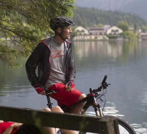 A woman lies on the park bench by the lake and next to her sits a man on his mountain bike